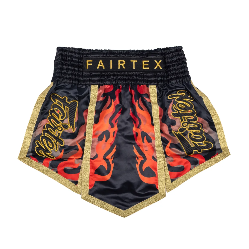 Мъжки шорти за муай тай Fairtex Aquila - Volcano, с черен сатенен фон и смели графики на пламъци в жълто и оранжево, със златни акценти и лога на Fairtex отстрани.